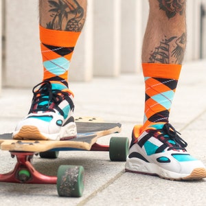Könnte beinhalten: Eine Person trägt orange-blaue gemusterte Socken mit schwarzen Sneakern und steht auf einem Longboard-Skateboard.