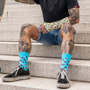 Op de afbeelding: Een man met een zonnebril en een zwart T-shirt zit op een betonnen trap met een longboard skateboard. Hij draagt ​​een blauwe jeans en blauwe en witte gepatroneerde sokken. Het skateboard heeft een blauw en wit ontwerp met het woord "Sunday" erop.