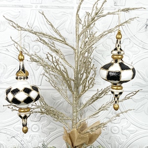 Puede incluir: Dos adornos navideños decorativos con un patrón de cuadros blancos y negros y detalles dorados cuelgan de un pequeño árbol dorado. Los adornos tienen una forma alargada única, añadiendo un toque de elegancia a la decoración navideña.