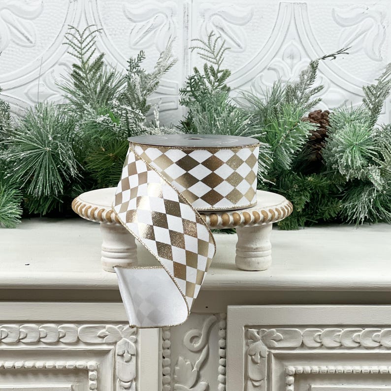 May include: A roll of white and gold harlequin patterned ribbon. The ribbon is displayed on a small, white, decorative stand. The background includes evergreen branches and a white, textured wall.