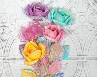 May include: A set of iridescent rose hair clips in pink, yellow, blue, and purple. The roses are made of plastic and have a shiny, pearlescent finish. The leaves are also iridescent and have a metallic sheen.