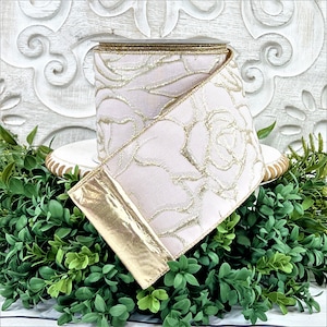A roll of light pink ribbon with gold embossed roses sits atop a white wwod pedestal surrounded by greenery. The end of the ribbon is turned back, revealing the reverse side, which is gold.