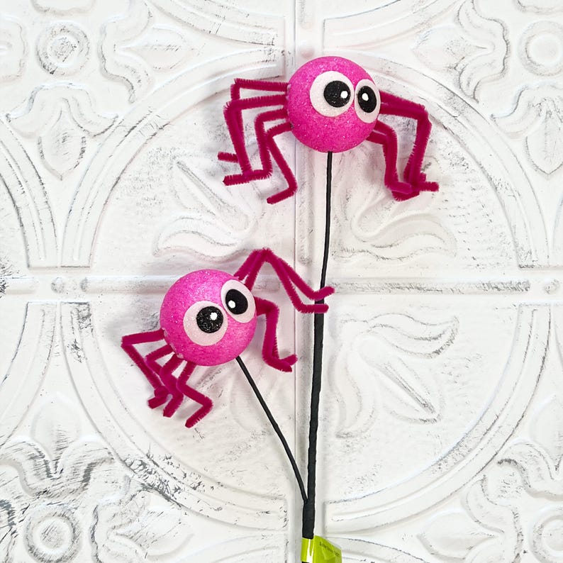 May include: Two pink glittery spider decorations with googly eyes and fuzzy legs. The spiders are attached to black stems, perfect for Halloween or spooky-themed crafts and decor.