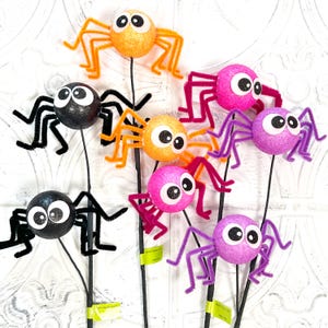 May include: A collection of colorful, glittery spider decorations on black stems. The spiders are orange, pink, purple, and black, with large googly eyes and pipe cleaner legs. These Halloween decorations are perfect for adding a spooky touch.
