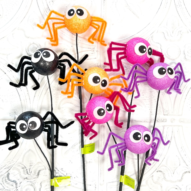 May include: A collection of colorful, glittery spider decorations on black stems. The spiders are orange, pink, purple, and black, with large googly eyes and pipe cleaner legs. These Halloween decorations are perfect for adding a spooky touch.