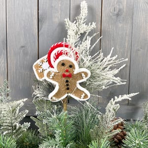 May include: A gingerbread man Christmas decoration with a red and white candy cane hat and a snowflake ornament. The decoration is surrounded by a snowy green pine tree branch.