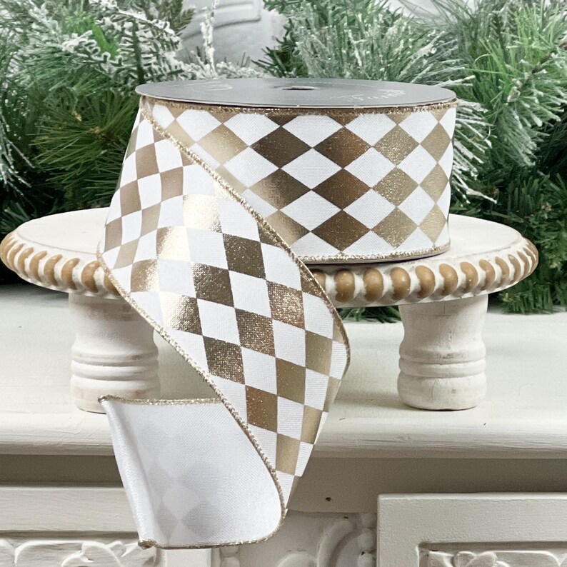 May include: A roll of white and gold harlequin patterned ribbon. The ribbon is displayed on a small, white, decorative pedestal. The ribbon has a metallic gold diamond pattern on a white background. The ribbon is suitable for gift wrapping and crafting.