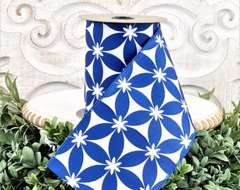 May include: A roll of blue and white floral patterned ribbon. The ribbon features a repeating pattern of white flowers on a blue background. The ribbon is displayed on a white wooden stand, surrounded by greenery.