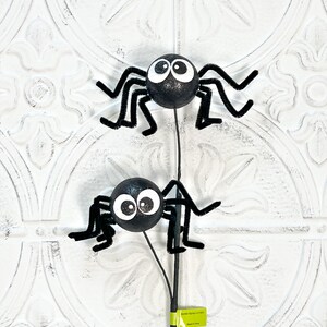 May include: Two black glitter spiders with large white eyes and pipe cleaner legs on a black stem. The spiders are a Halloween decoration, perfect for adding a spooky touch to any space. The background is a white, textured surface.