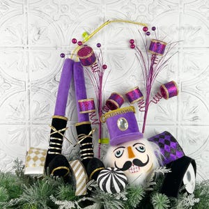 May include: A whimsical Christmas decoration featuring a nutcracker head with a black mustache and a purple hat. The decoration includes purple legs, decorative drums, and ribbons in gold, black, and white. The overall theme is festive and holiday-oriented.