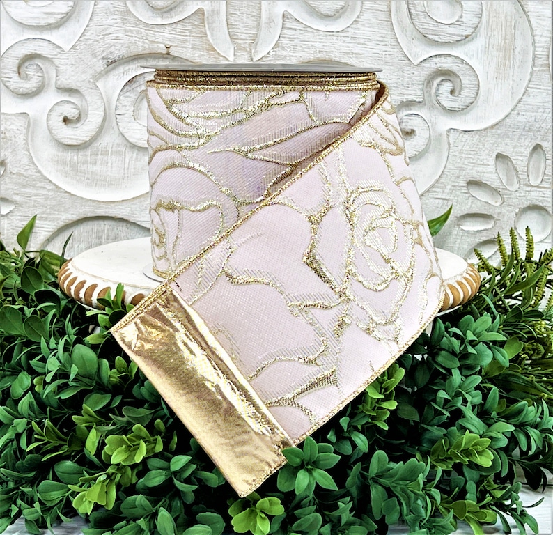 A roll of light pink ribbon with gold embossed roses sits atop a white wwod pedestal surrounded by greenery. The end of the ribbon is turned back, revealing the reverse side, which is gold.