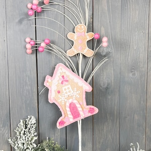 May include: A pink and brown gingerbread house and gingerbread person ornament with white glitter and pink balls on a white twig spray.