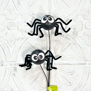 May include: Two black glitter spiders with large white eyes and pipe cleaner legs on a black stem. The spiders are a Halloween decoration, perfect for adding a spooky touch to any space. The background is a white, textured surface.