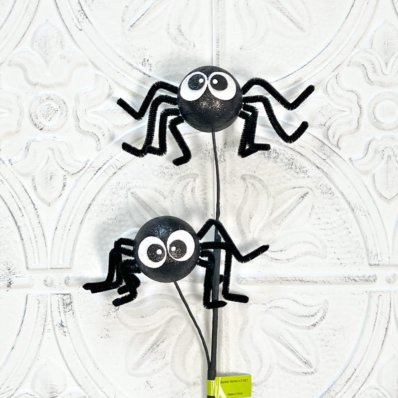 May include: Two black glitter spiders with large white eyes and pipe cleaner legs on a black stem. The spiders are a Halloween decoration, perfect for adding a spooky touch to any space. The background is a white, textured surface.