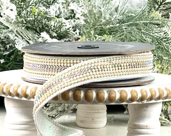 May include: A roll of white ribbon with a gold beaded trim and iridescent accents. The ribbon is displayed on a white, decorative stand with a beaded edge, against a backdrop of frosted greenery.