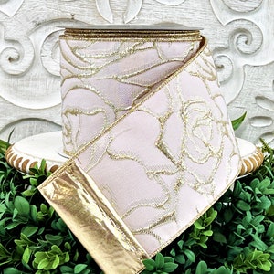 A roll of light pink ribbon with gold embossed roses sits atop a white wwod pedestal surrounded by greenery. The end of the ribbon is turned back, revealing the reverse side, which is gold.