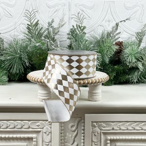 May include: A roll of white and gold harlequin patterned ribbon. The ribbon is displayed on a small, white, decorative stand. The background includes evergreen branches and a white, textured wall.
