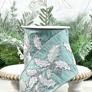 Puede incluir: Un rollo de cinta color turquesa con hojas y bayas de acebo bordadas en plata. La cinta tiene un acabado satinado y es ideal para manualidades y decoraciones navideñas. La cinta se exhibe sobre un soporte decorativo blanco y dorado.