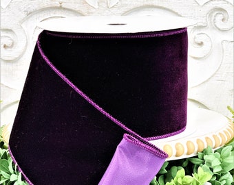 May include: A wide spool of deep purple velvet ribbon featuring a dark purple stitched edge. The ribbon is wound around a white cardboard core, showing a smooth, soft texture on the outer surface and a lighter lavender shade on the inner side.