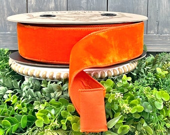 May include: A spool of bright orange velvet ribbon. The ribbon is thick and plush, with a rich, saturated colour. The spool is made of wood and sits on a decorative white and gold stand, surrounded by greenery.