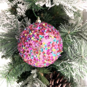 Peut inclure: Une boule de Noël rose, ornée de paillettes colorées et de petites perles, est suspendue à un sapin de Noël vert. La boule a un capuchon en métal doré et est entourée d'aiguilles de pin et de neige artificielle.