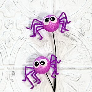 May include: Two decorative purple glitter spiders with googly eyes and pipe cleaner legs. The spiders are attached to a black stem, perfect for Halloween or spooky-themed crafts and decorations.
