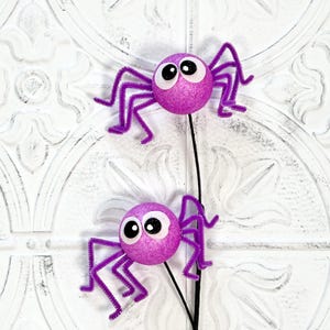May include: Two decorative purple glitter spiders with googly eyes and pipe cleaner legs. The spiders are attached to a black stem, perfect for Halloween or spooky-themed crafts and decorations.