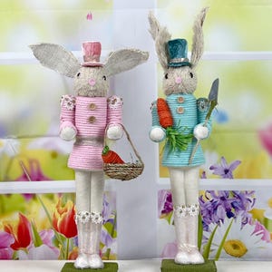 May include: Two Easter bunny figurines, one in pink and one in blue, with a carrot and a basket of carrots. The bunnies are dressed in a striped pattern and have a hat on their heads.