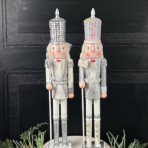 May include: Two decorative nutcrackers in a Christmas theme. One nutcracker has a silver sequined hat and the other has a pink sequined hat. Both have white hair, red mouths, and are dressed in silver and white uniforms. They are holding a staff and an axe.