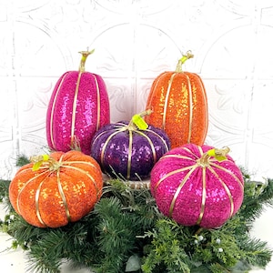 Puede incluir: Un conjunto de calabazas decorativas en varios tamaños y colores, incluyendo naranja, rosa y morado. Cada calabaza está cubierta de lentejuelas y adornada con ribetes y tallos dorados. Las calabazas están dispuestas sobre un lecho de follaje verde.