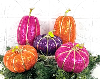 May include: A collection of decorative pumpkins in various sizes and colors, including orange, pink, and purple. Each pumpkin is covered in sequins and accented with gold-colored trim and stems. The pumpkins are arranged on a bed of green foliage.