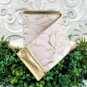 A roll of light pink ribbon with gold embossed roses sits atop a white wwod pedestal surrounded by greenery. The end of the ribbon is turned back, revealing the reverse side, which is gold.
