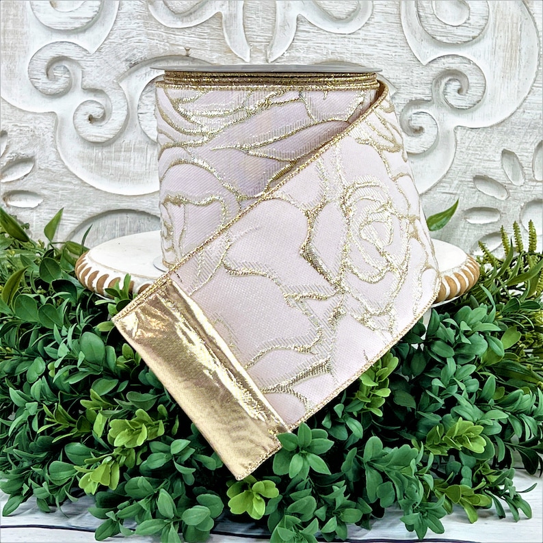 A roll of light pink ribbon with gold embossed roses sits atop a white wwod pedestal surrounded by greenery. The end of the ribbon is turned back, revealing the reverse side, which is gold.