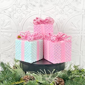 May include: Three decorative gift boxes in shades of pink and mint green, each adorned with a matching bow and ribbon. The boxes have a sequined texture, creating a shimmering effect. The arrangement is set against a white, patterned backdrop, with greenery at the base.