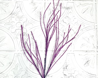 May include: A single stem of decorative purple glittered branches. The branches are thin and delicate, with a dark purple stem. The background is a white, textured surface with a repeating pattern.