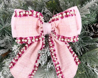 May include: A large, pale pink bow made of textured fabric, adorned with small pearls and dark pink detailing. The bow is set against a backdrop of frosted evergreen branches, suitable for festive occasions.