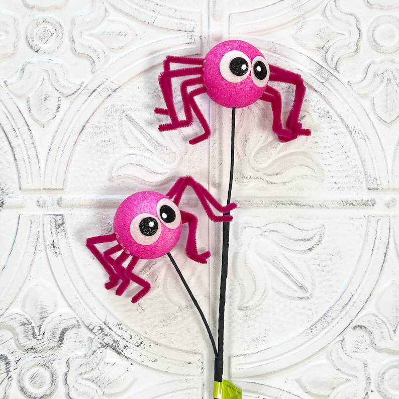 May include: Two pink glittery spider decorations with googly eyes and fuzzy legs. The spiders are attached to black stems, perfect for Halloween or spooky-themed crafts. The background is a white, textured surface.