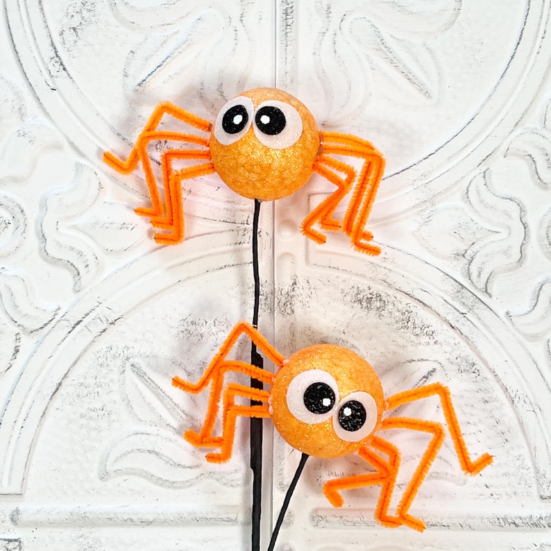 May include: Two orange glittery spider decorations with googly eyes and orange pipe cleaner legs. The spiders are attached to black stems, perfect for Halloween or spooky-themed decor.