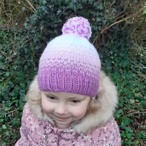 May include: A knitted hat with a gradient of white to purple, topped with a fluffy pom-pom. The hat is worn by a person wearing a pink floral jacket with a fur-lined hood. The background features green foliage.
