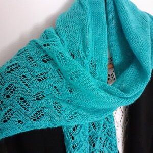 May include: A teal blue knitted scarf with a delicate lace pattern. The scarf is draped over a black jacket.
