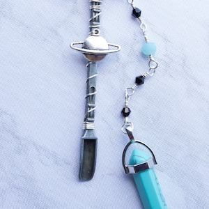 Op de afbeelding: Een zilveren ketting met een planeetbedel, zwarte kralen en een turquoise kristallen hanger.