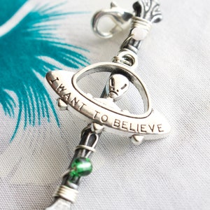 May include: Silver charm with a UFO design and the words "I want to believe" engraved on it. The charm is attached to a silver chain with green beads.