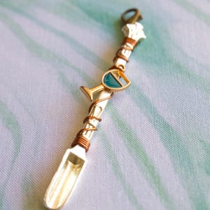 May include: A silver spoon with copper wire wrapped around it. A small gold charm with a green glass wine glass is attached to the wire. The spoon is on a white and green background.