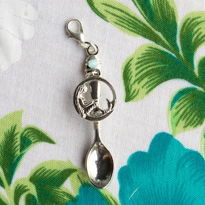 Puede incluir: Un charm de cuchara de plata con un diseño de sirena y una cuenta azul claro. El charm está unido a un cierre de plata.