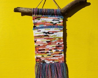Puede incluir: Un tapiz tejido de pared colorido con flecos, sujeto a una rama natural. El tapiz presenta un patrón geométrico en tonos de rosa, azul, verde, amarillo, blanco y negro.
