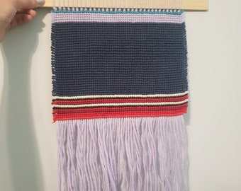Puede incluir: Un tapiz tejido para colgar en la pared con un centro azul y rayas rojas, blancas y negras. El tapiz tiene una franja de hilo de color lila claro.