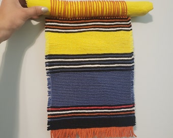 Puede incluir: Un tapiz tejido de pared colorido con un patrón de rayas amarillas, azules, negras, blancas y naranjas. El tapiz está sujeto a una rama pintada de amarillo con hilo naranja.