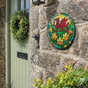 May include: A stained glass roundel depicting a red dragon and daffodils, mounted on a stone wall. A green door with a wreath and a black lantern are visible. A terracotta pot with daffodils sits to the left of the door, adding a touch of colour.