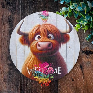 Puede incluir: Un letrero redondo de madera con una vaca Highland de dibujos animados con pelaje marr&oacute;n y cara blanca. La vaca est&aacute; mirando hacia arriba y tiene una expresi&oacute;n amigable. El letrero tiene las palabras "Bienvenido" y "Three Sisters Creations" impresas.