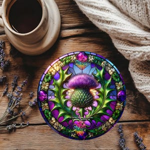 May include: A circular coaster with a stained-glass effect, showcasing a purple thistle, green leaves, and small white flowers. The coaster has a blue and purple border. The text "Three Sisters Creations" is visible on the coaster.